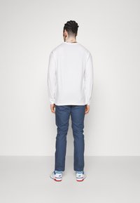 White long-sleeve shirt paired with blue trousers and white sneakers featuring blue and red accents, photographed from the back.