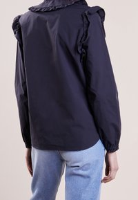 Blusa blu navy con dettagli arruffati sulle spalle, tessuto leggero e polsini elastici, abbinata a jeans azzurri.