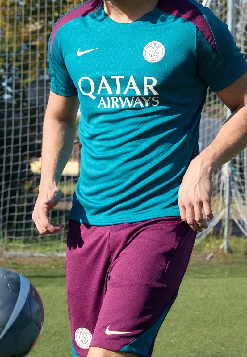 Teal urheilupaita, jossa on purppuranvärisiä yksityiskohtia, jossa on "QATAR AIRWAYS" -logo, yhdistettynä purppuranvärisiin shortseihin ja pyöreään joukkueen logoon lahkeessa.