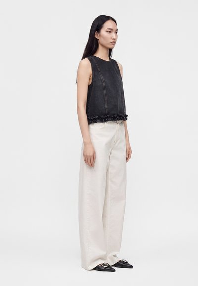 Femme aux longs cheveux foncés portant un haut noir sans manches avec des volants et un pantalon blanc large, debout devant un fond clair uni.