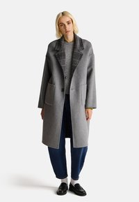 Cappotto grigio in lana con ampie tasche frontali, colletto a scacchi a contrasto e chiusura con un solo bottone, abbinato a jeans scuri e mocassini neri.