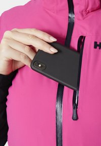 Růžová vesta s černým zipem a logem. Ruka pokládá černý kryt telefonu do levé kapsy se zipem. Hladký povrch.