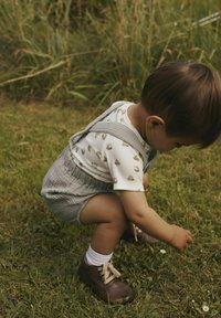 Barn hukande på gräs, bär en ljus randig skjorta med båt mönster och gråa shorts, bruna skor och vita strumpor.