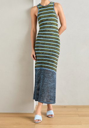 Woman in sleeveless, fitted striped dress with blue mesh hem, standing against a light wall, wearing light blue open-toe sandals.