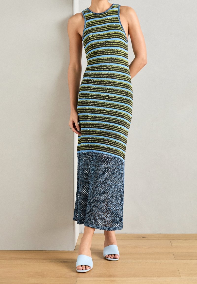 Femme en robe rayée ajustée sans manches avec ourlet en mesh bleu, debout contre un mur clair, portant des sandales bleu clair à bout ouvert.