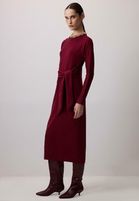 Burgundy hosszú ujjú ruha, deréknál kötőszalaggal, amelyet azonos színű, térdig érő, hegyes orrú csizmával párosítottak. Simább szövet és testhez simuló fazon.