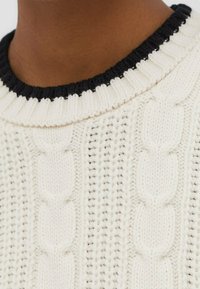 Pull crème en maille torsadée avec un motif texturé et un col côtelé noir. Le tissu semble doux et épais.
