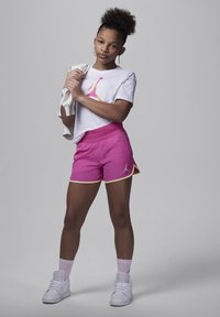 Pantaloni corti rosa con rifiniture gialle, t-shirt bianca con un logo colorato, sneakers bianche e calze rosa chiaro, in piedi contro uno sfondo grigio.