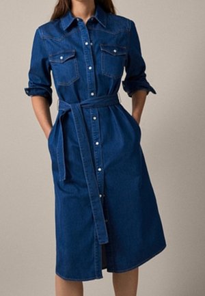 Woman wearing a mid-length dark blue denim shirt dress with rolled-up sleeves, button closure, chest pockets, and a waist belt.