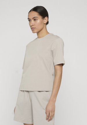 Sötét haját hátrafogva viselő nő, bézs színű rövid ujjú pólót és hozzáillő rövidnadrágot visel, egyszínű világos háttér előtt áll.