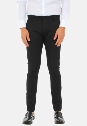 Uomo che indossa pantaloni neri aderenti, camicia bianca con bottoni e mocassini neri in pelle, in piedi davanti a uno sfondo bianco uniforme.