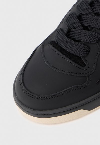 Nahaufnahme der Zehenpartie eines schwarzen Sneakers mit schwarzer Naht, dicken schwarzen Schnürsenkeln und einer beigen Gummisohle auf einem weißen Hintergrund.