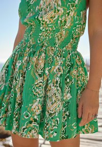 Zielona sukienka z marszczonym stanem i rozkloszowanym dołem, zdobiona odważnym złoto-białym wzorem kwiatowym na teksturowanej tkaninie.