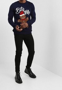 Pull en maille bleu marine avec le texte "Mords-moi" et un design de bonhomme en pain d'épice. Un pantalon noir slim-fit et des bottines à lacets noires complètent la tenue.