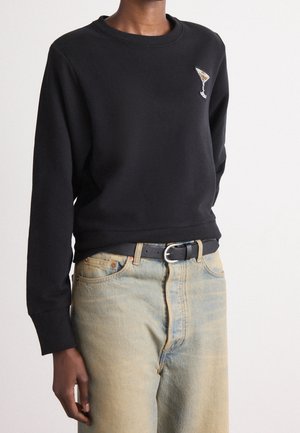 Personne portant un sweat-shirt à col rond noir avec un petit verre à martini brodé sur la poitrine, un jean en denim bleu clair et une ceinture noire.