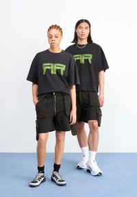 Duas modelos vestem camisetas pretas oversized combinando, com letras gráficas verdes, acompanhadas de calções cargo pretos com zíperes e bolsos.