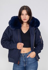 Marineblau bomberjacke mit großem Kunstfellkragen, Reißverschluss, gerippten Bündchen und taillierter Passform. Kombiniert mit einem schwarzen Oberteil und Jeans.