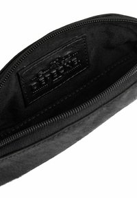 Primer plano de un bolsillo negro con cremallera abierto que muestra una etiqueta interior cosida con el nombre de la marca en relieve: Detective.
