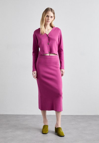Fuchsia kurzgeschnittene Strickjacke mit Knöpfen, kombiniert mit einem fuchsia-farbenen knielangen Rock und olivgrünen klobigen Clogs mit Absatz. Glatte Strickstruktur.