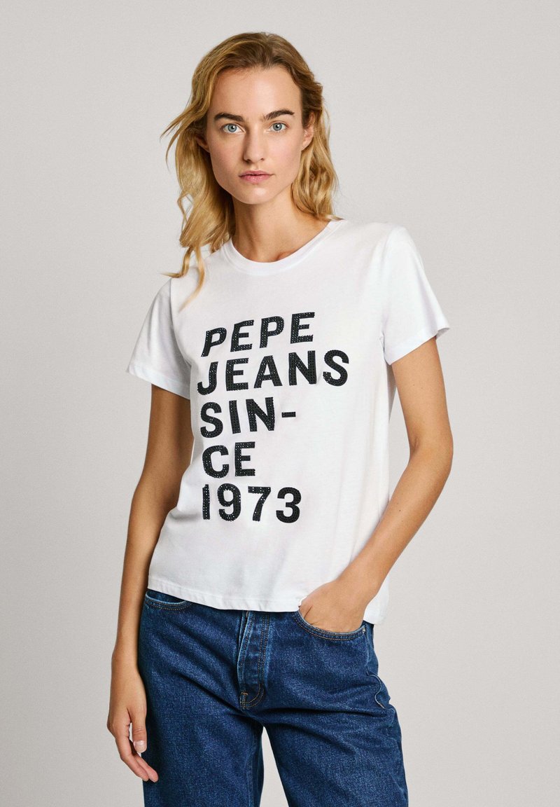 Camiseta blanca de algodón con un atrevido texto negro que dice "PEPE JEANS SINCE 1973". Manga corta y un corte relajado, combinada con unos pantalones vaqueros de denim azul oscuro.