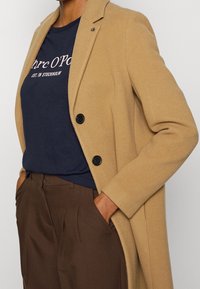 Kamelový kabát s přiléhavým střihem, zdobený černými knoflíky a hladkým povrchem, přeložený přes námořnické tričko s bílým nápisem.