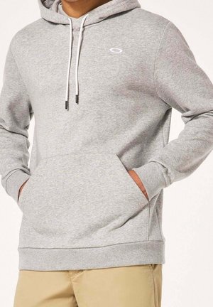 Homme portant un sweat à capuche gris clair avec poche avant et cordons blancs, avec un pantalon beige, mains dans les poches.