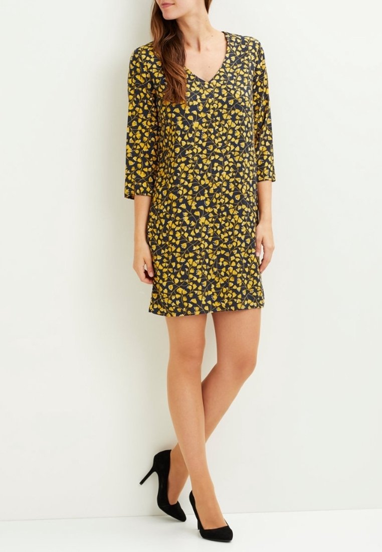 Femme portant une robe noire et jaune à motifs, longueur genou, manches trois-quarts et talons noirs, debout sur un fond blanc.