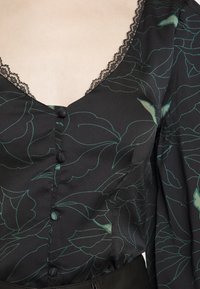 Blouse noire avec motif de feuilles vertes, encolure en V bordée de dentelle, boutons recouverts de tissu sur le devant et manches longues bouffantes.