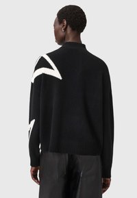 Czarny sweter robiony na drutach z wysokim kołnierzem, z białymi kątowymi wzorami na rękawach i ściągaczem na dole. Miękka struktura i luźny krój.