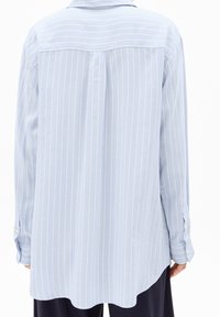 Vue arrière d'une personne portant une chemise à manches longues rayée verticalement bleu clair avec des poignets boutonnés et un pantalon large sombre.