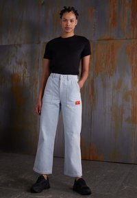 T-shirt noir à manches courtes, jean large taille haute bleu clair avec une étiquette rouge, et baskets noires. Fond texturé avec des détails rouille.