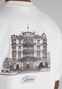 White t-shirt featuring a black line drawing of a grand building with a detailed entrance and trees, labeled "Prohibited Grand Resort."