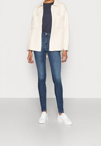 Chaqueta blanca texturizada con dos bolsillos frontales, llevada sobre una camisa azul marino y combinada con jeans ajustados azules y zapatillas blancas.