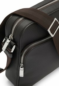 Sort læder crossbody taske med struktureret overflade, sølv lynlåse og justerbar brun strop. Flere rum til organisering.