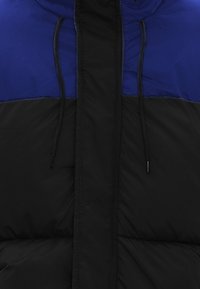 Geacă pufoasă cu o secțiune superioară albastră și o secțiune inferioară neagră. Textură groasă, quilted, cu șnururi reglabile la guler.