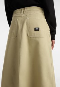 Beige kjol med en slät textur, överdimensionerade fickor, vit sömnad och en svart Vans-logotagg på baksidan.