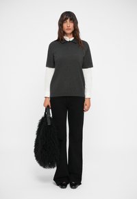 Polo shirt grigio scuro a maniche corte sopra una camicia bianca, abbinato a pantaloni neri a zampa d'elefante e con una borsa nera e soffice. 