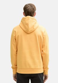 Gelbes Hoodie mit tailliertem Design, großer Kapuze, gerippten Bündchen und Saum. Glatte Textur, getragen mit dunklen Hosen, von hinten gesehen.