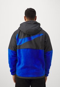 Hættejakke med en sort overdel og en livlig blå fleece underdel. Har et fremtrædende Nike-logo og elastiske manchetter.