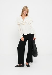 Witte, rimpelige blouse met knopen aan de voorkant, gecombineerd met zwarte wijde broek. Model houdt een zwarte tas vast, met platte zwarte schoenen. Schone achtergrond.