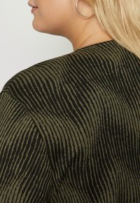 Top z bavlněné směsi s proužkovaným vzorem v černé a olivově zelené barvě, hladkou texturou a volným střihem, zaměřeno na pohled zezadu.