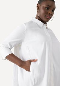 Camicia bianca a maniche lunghe con colletto button-down, caratterizzata da una vestibilità rilassata e tasche laterali. Texture morbida e tessuto liscio.