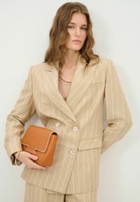 Beige stribet blazer med dobbelte knapper og lomme, parret med en beige top. Holder en brun lædertaske med dekorerende syninger.
