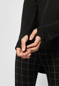 Černá mikina na zip s dlouhými rukávy a otvory na palce, vyrobena z jemné látky. Nosí se s černými legínami s kostkovaným vzorem.