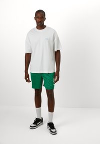 White oversized t-shirt with light blue text, paired with green patterned shorts. Includes black shoes and white knee-high socks.