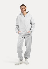 Szary hoodie i dopasowane dresy wykonane z miękkiej mieszanki bawełny. Posiada kieszenie, zamek błyskawiczny i elastyczne mankiety. Noszone z białymi sneakerami.