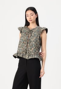 Braune Bluse mit floralem Muster, kurzen Ärmeln, V-Ausschnitt, Frontbindebändern und einem rüschierten Saum, kombiniert mit schwarzen Hosen.