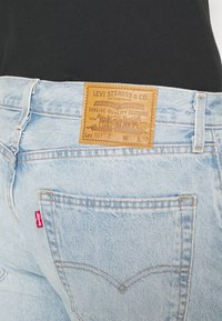 Svijetloplave traperice s kožnom oznakom koja sadrži brendiranje "Levi Strauss & Co.". Kontrastni žuti šavovi i crvena oznaka na stražnjem džepu.