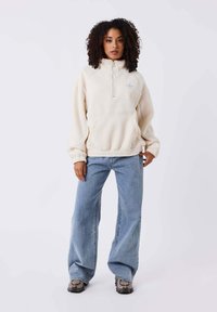 Pullover in pile color crema con colletto alto e mezza zip, dotato di una tasca frontale. Abbinato a jeans in denim blu larghi e scarpe a fantasia.