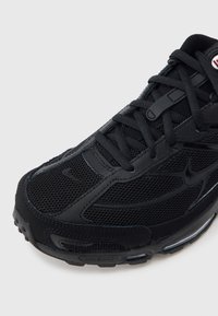 Čierna športová topánka s priedušnou sieťovinou a syntetickými prekrytiami, s zaoblenou špičkou, textúrovanou tkaninou a šnúrkami. Na jazyku viditeľné logá značky.